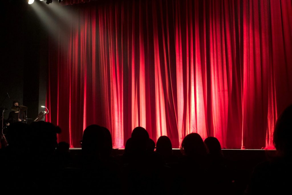 Home Dimly lit theater stage with red curtains and audience silhouettes under spotlights.