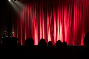 Dimly lit theater stage with red curtains and audience silhouettes under spotlights.