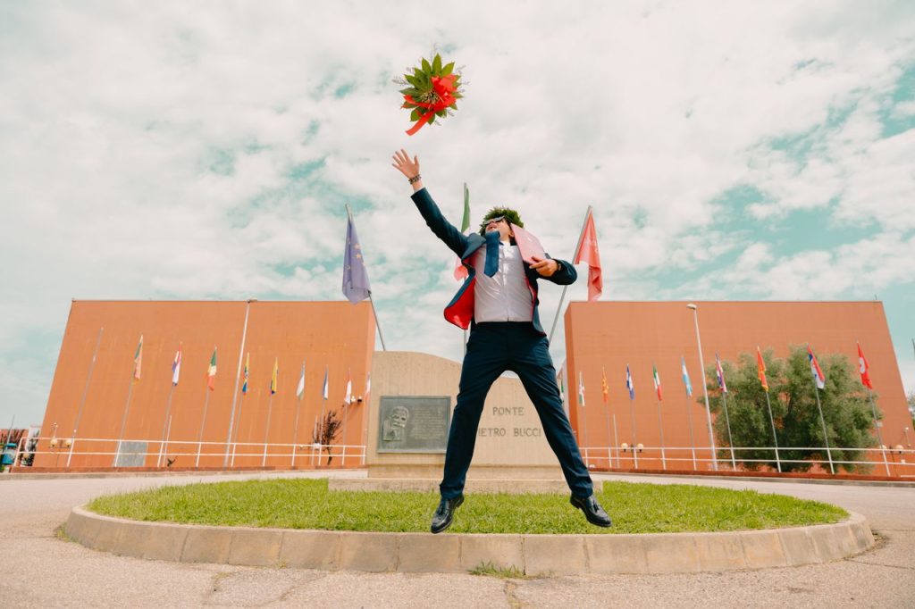 Young graduate throwing bouquet in front of modern building, celebrating success in an urban setting.